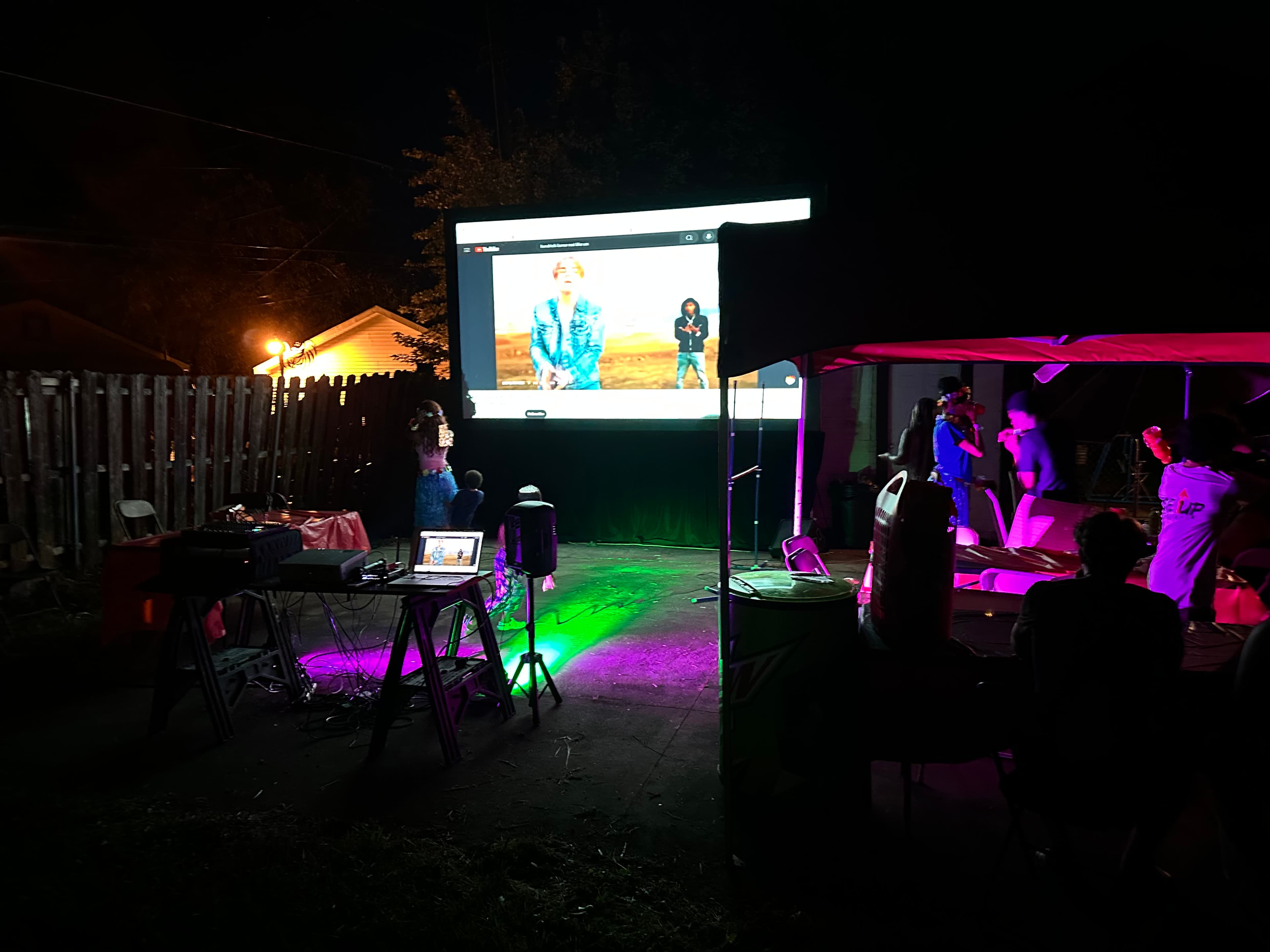 An outdoor evening event lit with colored uplighting beside a projection screen